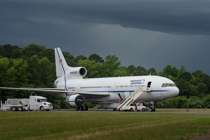 A white plane on a tarmac next to a truck and car, with blue 'Northrop Grumman' livery.