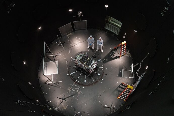 Two men standing in a circular room in white protective clothing, next to a bulky machine, ladders and screens.
