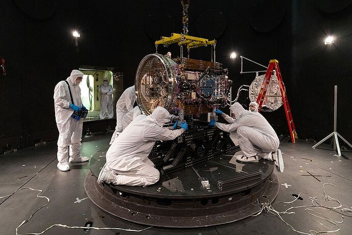Five people in protective white clothing bending around a bulky machine with a domed ending, in a dark open area.