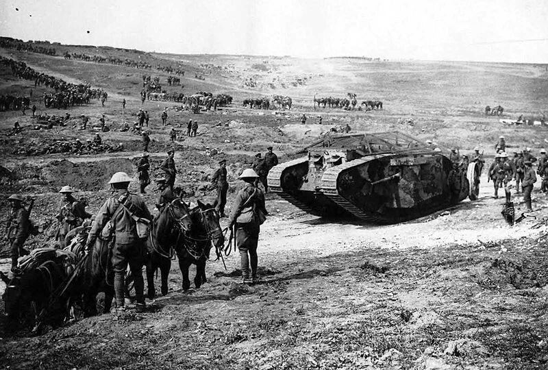 British Mark I tank, apparently painted in camouflage, flanked by infantry soldiers, mules and horses.