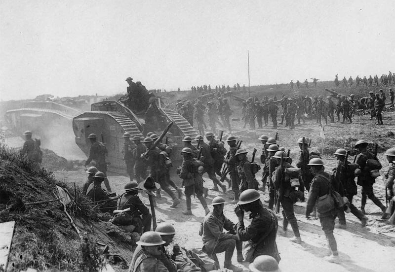 Allied advance on Bapaume, France, ca. 1917. Two tanks are moving towards the left, followed by troops. In the foreground some soldiers are sitting and standing at the roadside. One of them appears to be having a drink. Beside the men is what appears to be a rough wooden cross with an Australian or New Zealand service hat on it. In the background other troops are advancing, moving field guns and mortars.